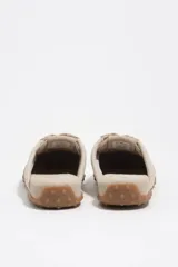 Zapatilla estilo mula de color beige, confeccionada en una combinación de algodón y serraje vacuno, con suela de caucho con tachuelas texturizadas y costuras decorativas visibles en la parte superior.
