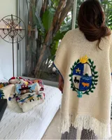 Ruana de lana merino color crema, tejida y bordada a mano, con el escudo de Uruguay en la espalda y flecos en el ruedo.