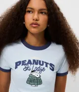 Remera de algodón color celeste con cuello y bordes de mangas en color azul marino. Presenta estampa frontal con el personaje Snoopy vestido con campera puffer y la inscripción Peanuts Ski Lodge.