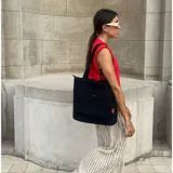 Cartera tipo shopper de lona negra, con gran compartimento principal, doble asa de hombro y bolsillo frontal con solapa y botón a presión.