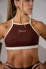 Top deportivo de cuello halter en color marrón chocolate con ribetes en color crema. Presenta el logo de la marca estampado en el centro del pecho.
