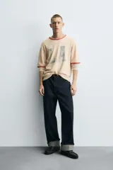 Camiseta de punto tejida color beige claro, corte boxy fit, con cuello redondo y mangas cortas. Presenta estampado desgastado con las letras "LA" en el frente y ribetes en color terracota en cuello y mangas. El bajo es irregular.