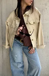 Sobrecamisa beige de gabardina con tachas metálicas decorativas en el cuello y la solapa frontal. Cuenta con bolsillos con solapa en el pecho y cierre frontal con botones.