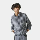 Camisa de franela a cuadros en tonos grises, negros y azules, con cuello clásico, corte ligeramente oversize, cierre frontal con botones y bolsillo en el pecho con logo bordado.