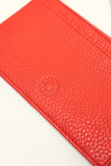 Monedero rectangular de cuero vacuno con textura granulada en color marrón topo. Cuenta con cierre superior metálico y un tirador decorativo de cuero en color naranja con un nudo. Presenta un pequeño logo grabado en el centro.
