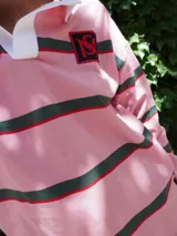 Buzo tipo rugby de algodón con diseño de franjas horizontales en tonos beige, negro y fucsia. Presenta cuello polo en contraste y cierre frontal parcial.