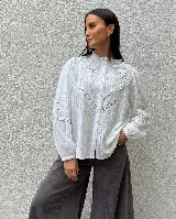 Camisa blanca de estilo romántico con detalles de encaje y alforzas en el frente. Presenta cuello alto con volado, mangas largas con puños elásticos y cierre frontal de botones.