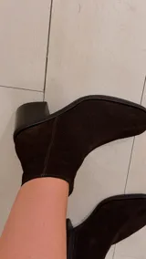 Bota de caña alta de gamuza en color marrón chocolate, con taco cuadrado y puntera cuadrada. Cuenta con un diseño holgado en la caña y detalle de trabilla decorativa en el tobillo.