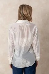 Camisa de manga larga en color blanco, confeccionada en tejido traslúcido con delicados bordados florales en hilo de seda al tono. Presenta cuello clásico, cierre frontal con botones y puños abotonados.