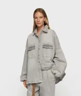 Camisa oversize de algodón orgánico con certificación GOTS, color gris claro con efecto lavado. Presenta cuello, cierre frontal de botones, dos bolsillos de parche en el pecho y puños elásticos.