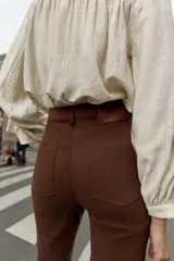 Pantalón de tiro alto color marrón oscuro, con corte flare (campana) y nervio marcado en el frente de ambas piernas. Presenta bolsillos delanteros y bolsillos de plastrón en la espalda.