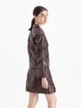 Chaqueta de cuero vacuno color marrón con acabado brillante, diseño de cuadros con líneas finas en tonos rojizos y beige, y detalle de volados de cuero aplicados en forma de trama. Posee cuello camisero, cierre frontal con botones y mangas largas.