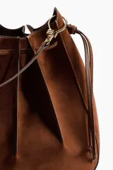 Cartera bandolera de gamuza color marrón, con diseño de pliegues cosidos y cordón ajustable en la parte superior. Cuenta con correa de hombro ajustable y herrajes metálicos.