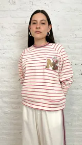 Buzo blanco a rayas horizontales rosas con cuello redondo a rayas rosas, marrones y blancas. Tiene un parche con la letra M dorada y una flor rosa.