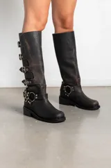 Botas de caña alta confeccionadas en cuero con efecto gastado. Presentan un diseño de estilo biker con múltiples hebillas laterales y apliques metálicos, incluyendo una argolla decorativa en el tobillo. Cuentan con un taco bajo cuadrado y suela de goma.
