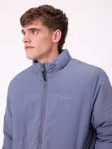 Campera reversible de nylon con cuello alto y cierre frontal. Un lado es de color gris liso con logo bordado, mientras que el otro lado presenta un estampado abstracto en tonos azules. Cuenta con bolsillos laterales y puños elásticos.
