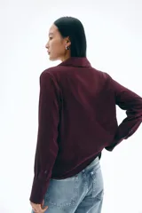 Blusa de manga larga color borgoña, con cuello clásico y escote en V con botones frontales. La tela tiene un acabado satinado y el bajo es redondeado.