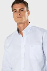 Camisa Oxford lisa de color celeste, con cuello abotonado, manga larga, cierre frontal de botones y un bolsillo aplicado en el pecho con detalle bordado.