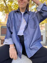 Sobre camisa de gabardina símil denim, color azul, con costuras visibles en color rojo y etiqueta de cuero en la espalda. Presenta un corte holgado y mangas largas.