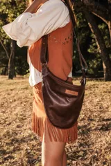 Bandolera de cuero 100% natural, color marrón oscuro, con diseño minimalista y acabado artesanal. Tiene una correa ajustable y un parche con el logo de la marca.