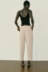 Pantalón tobillero de tiro medio, color beige, con pinzas delanteras y bolsillos frontales. Incluye un cinturón fino de cuero marrón con hebilla metálica dorada y cierre frontal con cremallera, botón interior y gancho.