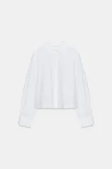 Camisa blanca de popelín con cuello solapa, mangas largas abullonadas con puño y bajo recto. Cierre frontal con botones ocultos por solapa.