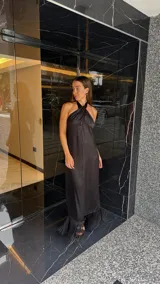 Vestido largo negro con escote halter cruzado y abertura frontal. El ruedo presenta un detalle de flecos.