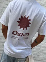 Remera blanca de corte oversize con cuello redondo y manga corta. Presenta un estampado frontal en color marrón rojizo con un diseño abstracto que incluye un sol estilizado y la palabra "Osten".