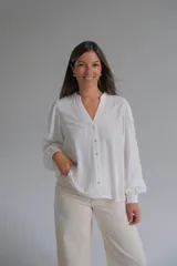 Blusa blanca de corte holgado con cuello en V, cierre frontal de botones y mangas largas abullonadas con puños elásticos. Presenta detalles bordados sutiles en la parte frontal y en las mangas.