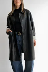 Trench de ecocuero negro de corte relajado y largo medio. Presenta cierre frontal con botones, bolsillos delanteros funcionales y tajo posterior. Las mangas cuentan con un forro interno a rayas que queda visible al doblarlas.