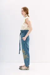 Pantalón de denim azul de tiro alto con cintura estilo paper bag y lazo a tono. Presenta un bordado elaborado con lentejuelas y piedras en forma de palmeras en la parte lateral de la pierna.