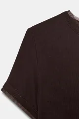 Camiseta de manga corta color marrón oscuro con textura acanalada, cuello redondo y bajo recto. Presenta un bajo superpuesto de tul o tela más clara, visible en el ruedo y en el cuello.