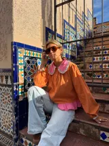 Camisa de manga larga color naranja con cuello grande en contraste color rosa y detalle de volados en el borde. Presenta un diseño con botones frontales y un detalle de tela rosa en la parte inferior.