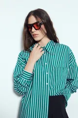 Camisa de silueta amplia y caída fluida, color celeste con rayas verticales blancas. Presenta cuello camisero clásico, cierre frontal con botones y un bolsillo aplicado en el pecho. Las costuras están resaltadas en azul oscuro.