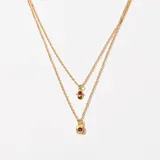 Collar doble dorado con cadena de eslabones finos. El collar superior tiene un dije de mano de Fátima con una piedra roja engarzada. El collar inferior tiene un dije de candado con una piedra roja engarzada.