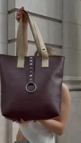 Bolso tote color borgoña con textura símil piel de cocodrilo, doble asa corta color beige y cierre superior. Presenta una tira vertical con apliques metálicos y un aro plateado decorativo.