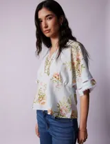 Blusa blanca estampada con flores azules, escote en V y mangas cortas amplias. Detalles de puntilla en escote y mangas.
