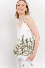 Blusa blanca de algodón liviano con tirantes finos y escote en V. Presenta un corte suelto con pechera fruncida y bordados artesanales en tonos verde y beige con motivos geométricos y florales.