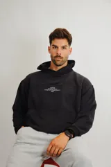 Hoodie negro de corte oversize con capucha, confeccionado en algodón. Presenta un diseño minimalista con un pequeño estampado tipográfico en el pecho que incluye el nombre de la marca y detalles de diseño.