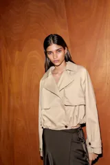 Trench corto de color beige con cuello de solapa y escote en pico. Presenta un diseño de manga larga con trabillas en los puños, bolsillos laterales y un bajo con acabado abullonado gracias a un elástico interior. Cuenta con cierre frontal cruzado con botones y forro interior.