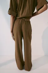 Pantalón de tiro medio color marrón, con cintura elástica y cordón ajustable con abalorios decorativos. Presenta bolsillos laterales y perneras rectas con diseño texturizado.