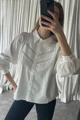 Camisa blanca de algodón con cuello clásico, cierre frontal de botones y mangas largas abullonadas con puños abotonados. Presenta un diseño con alforzas y detalles de encaje calado en el frente.