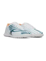 Championes de fútbol Under Armour Shadow Select 4 Turf, diseñados para césped artificial. Presentan un diseño aerodinámico en color blanco con detalles en azul petróleo y acentos en naranja. Cuentan con empeine sintético texturizado para mejor control, mediasuela Charged Cushioning para amortiguación reactiva y suela de goma con tacos multidireccionales.