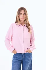 Camisa blanca de manga larga con cuello clásico y cierre frontal de botones negros en contraste. Presenta un bolsillo frontal en el pecho y un diseño de líneas simples y corte recto.