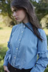 Camisa de jean azul claro con cuello alto, cierre frontal de botones y mangas largas con detalles de volados horizontales y puños con volados.