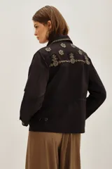 Campera estilo militar o safari color verde oliva, confeccionada en algodón. Presenta cuello solapa y bolsillos frontales con tapa. Destaca por sus apliques de pedrería o bordados en tonos marrones y dorados en el cuello y sobre los bolsillos.