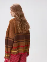 Pullover de punto con textura aterciopelada, diseño oversize, cuello polo con cierre frontal parcial y detalles de pespuntes en contraste. Presenta rayas horizontales en tonos tierra y un parche bordado con la letra M en el pecho.