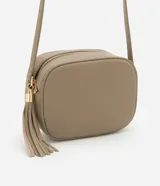 Cartera bandolera pequeña color beige, confeccionada en poliuretano con textura granulada. Presenta cierre superior con cremallera, correa larga ajustable y detalle de borla decorativa en el lateral con herrajes dorados.