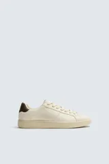 Zapatilla deportiva de diseño minimalista en color crema, con detalle de contraste en color marrón oscuro en el talón y suela de goma con volumen.
