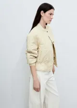 Chaqueta acolchada color crema, estilo bomber, con cuello perkins, cierre frontal con botones, dos bolsillos laterales y detalle fruncido en el bajo.